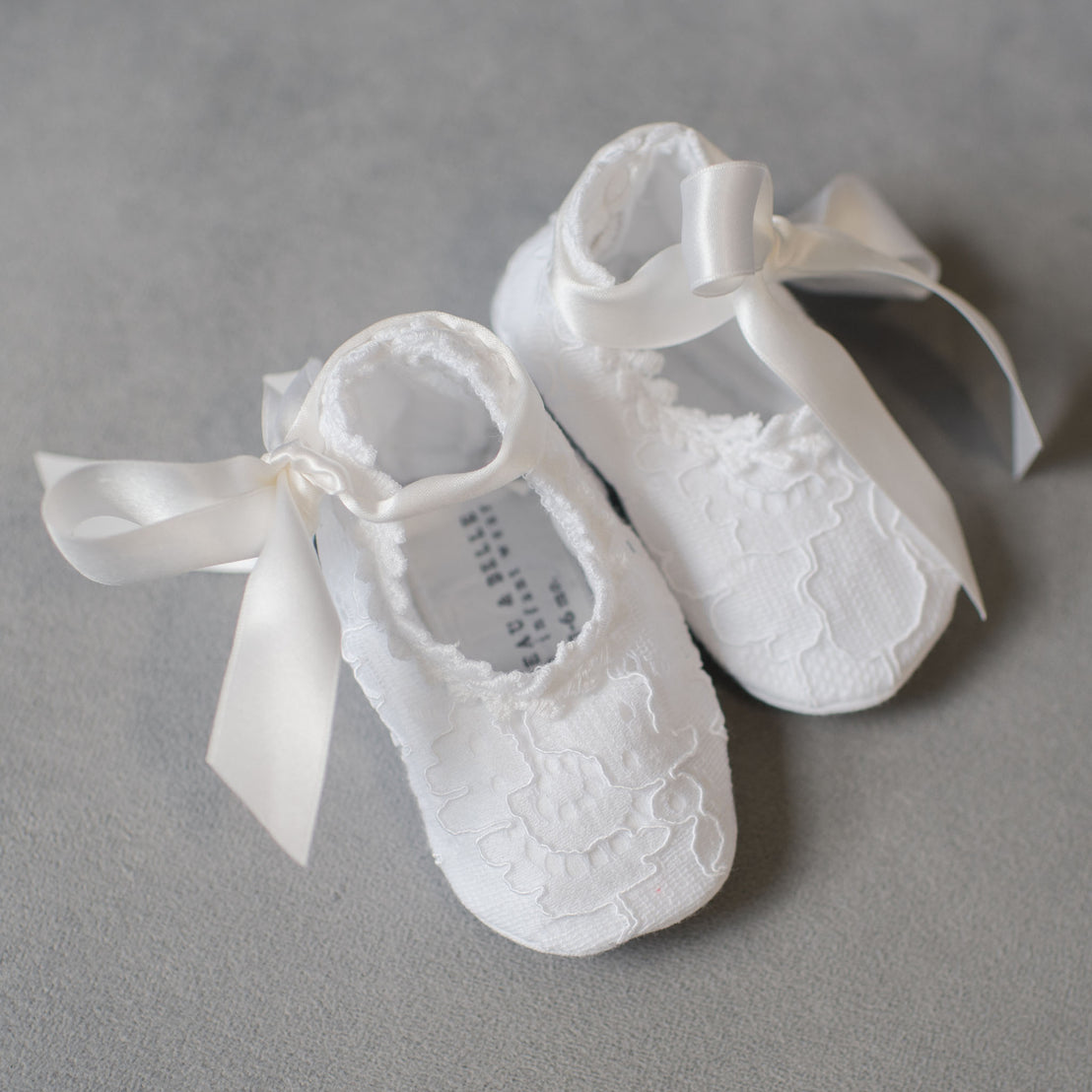 The Aria Lace Christening Booties laying on a soft grey surface. The Aria Lace Christening Booties made with white embroidered lace, lined with soft pima cotton, and fitted with white silk ribbon ties.