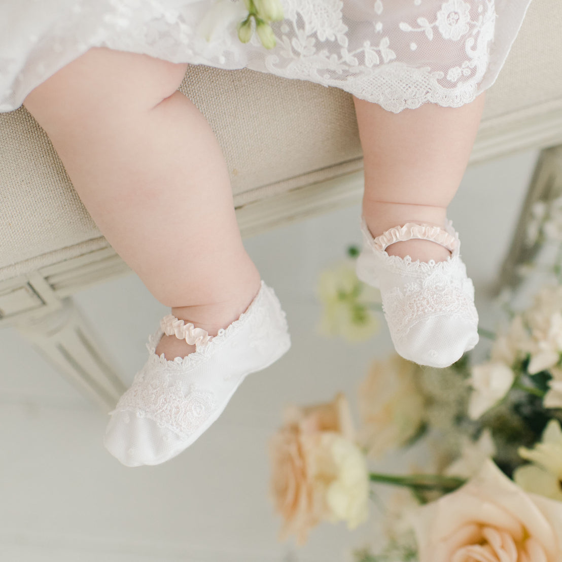 A close-up of a baby's feet adorned with the Melissa baby booties, hovering above a decorative arrangement of soft peach and cream flowers.