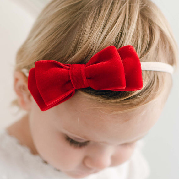 A close-up of a young child with an Emily Red Velvet Bow Headband, looking downward with their face partially visible at an upscale baptism. The background is soft and neutral.