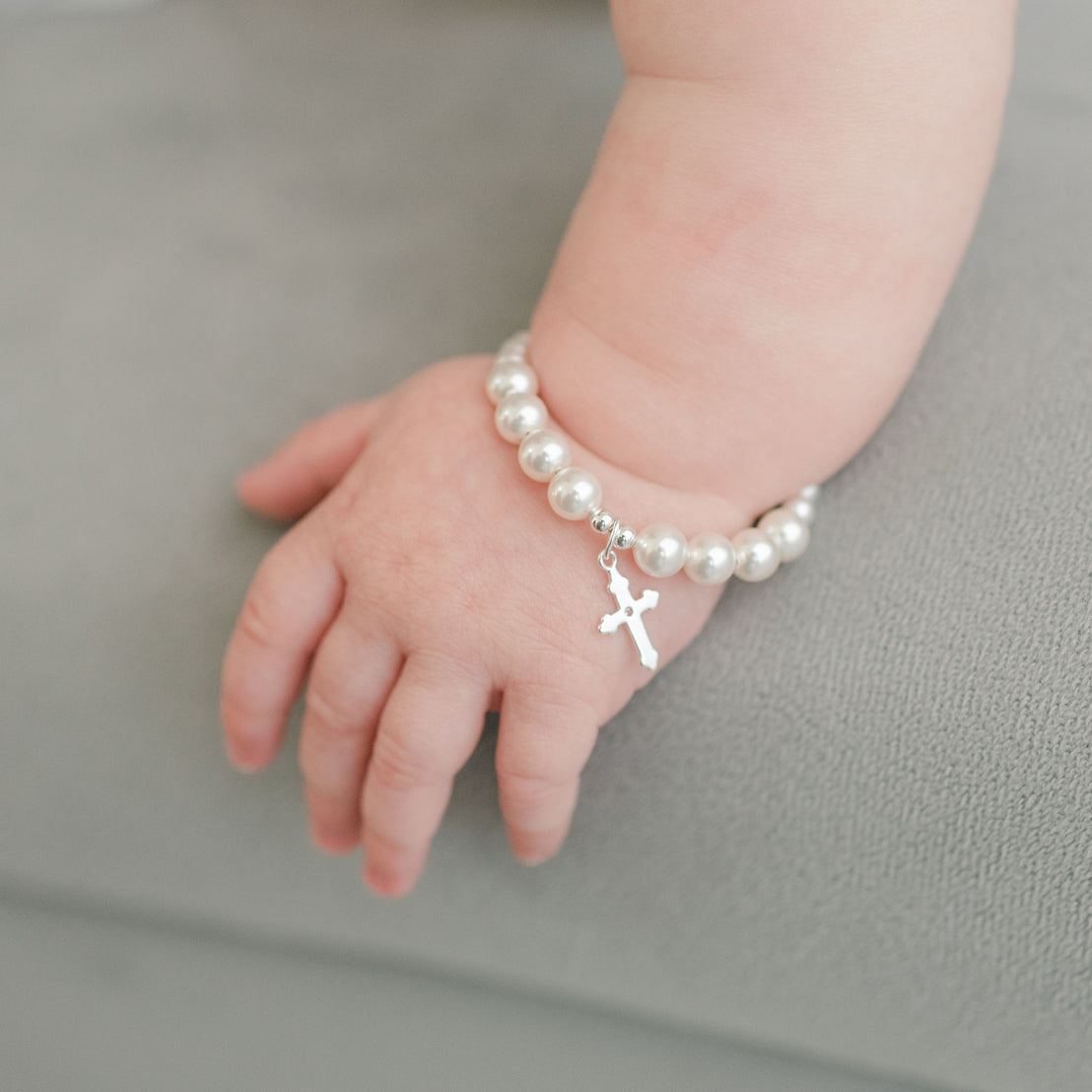 A close-up photo of a baby's wrist adorned with a White Luster Pearl Bracelet with Silver Cross, resting on a soft gray surface.