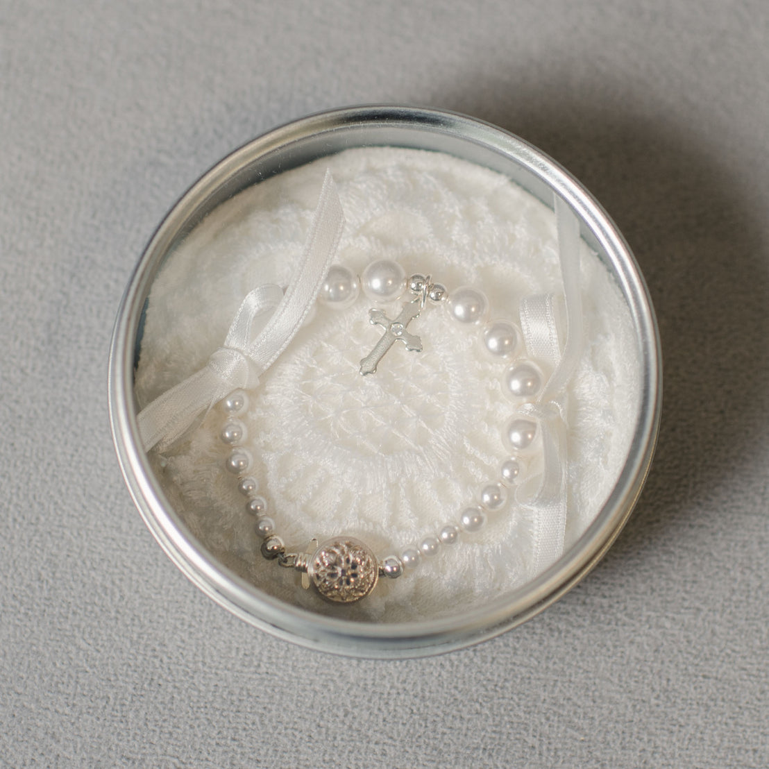 A White Luster Pearl Bracelet with Silver Cross charm and a circular clasp, laid on a white cushion inside a silver round box.