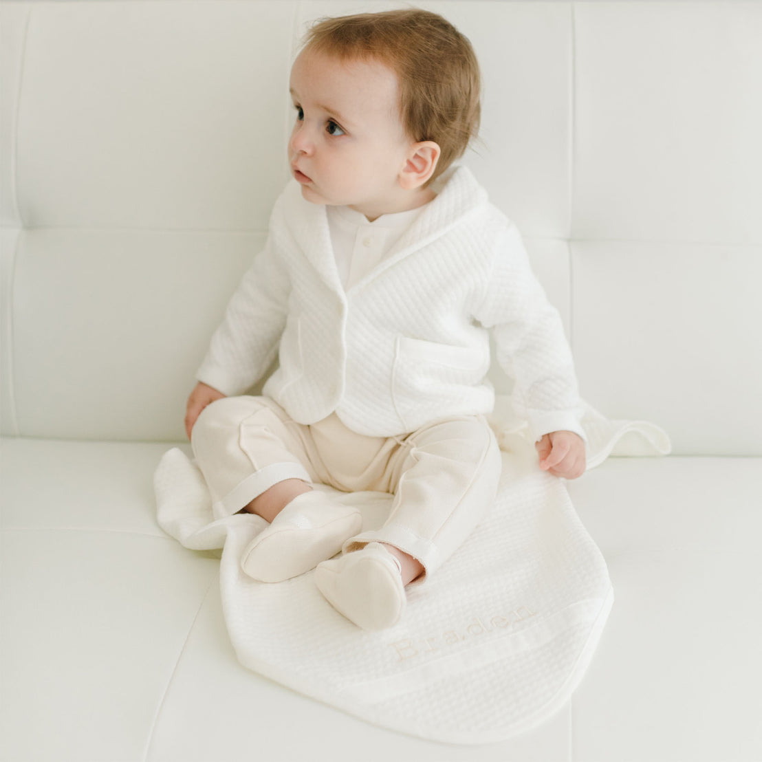 Baby boy sitting on a white couch with a white quilt blanket. He is wearing the Braden 3-Piece Suit
