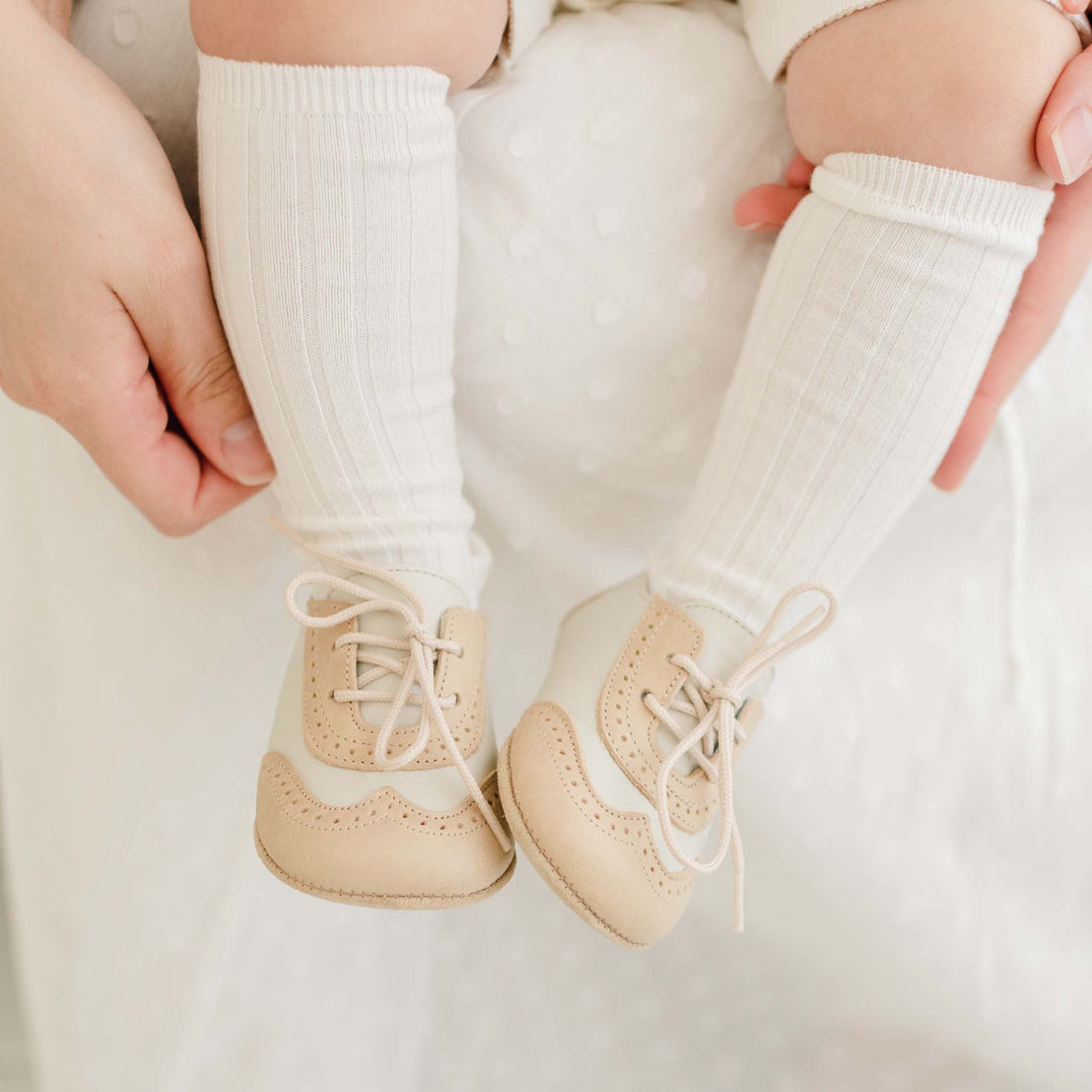 Baby wearing a ribbed knee-high socks and a pair of the Beige and Ivory Wingtip Shoes made with a tan and ivory matte leather with tan suede soles and detailed edging