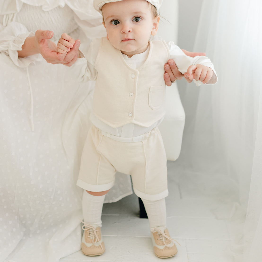 Baby boy standing up with the help of his mother. He is wearing the Braden Vest Shorts Suit. The vest and pants are made of ivory French Terry Cotton.