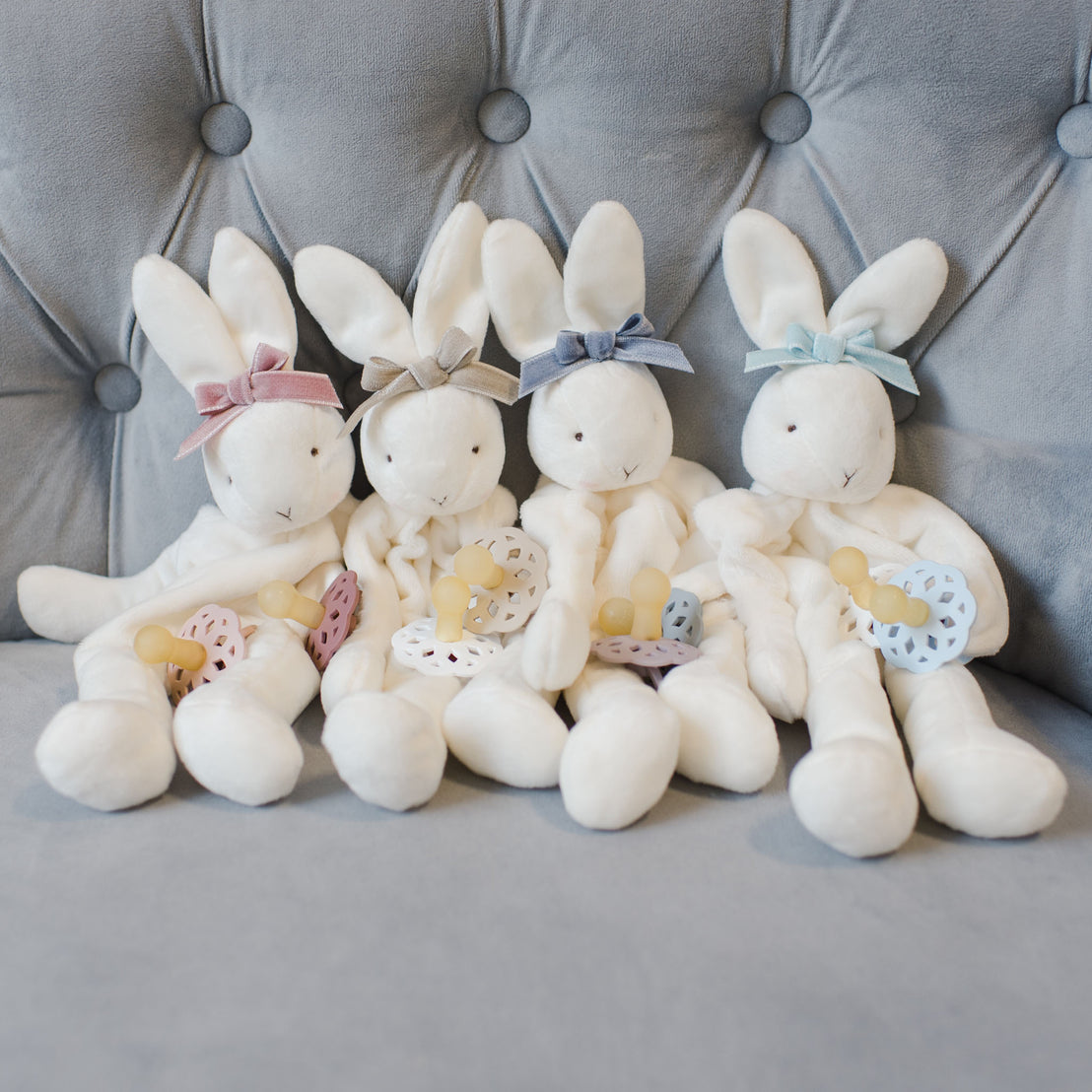 Flat lay photo of the Emily Silly Bunny Buddy and the color variations, including mauve, tan, heather, and sky blue. Made out of a soft white velour with a rattling head and hand loop to tie on to a pacifier.