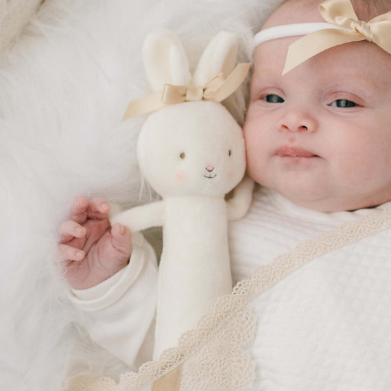 A baby with a gentle expression and wearing the Mia Bow Headband holds a Mia Bunny Chime Rattle, wrapped snugly in the Mia Personalized Blanket with delicate lace trim.