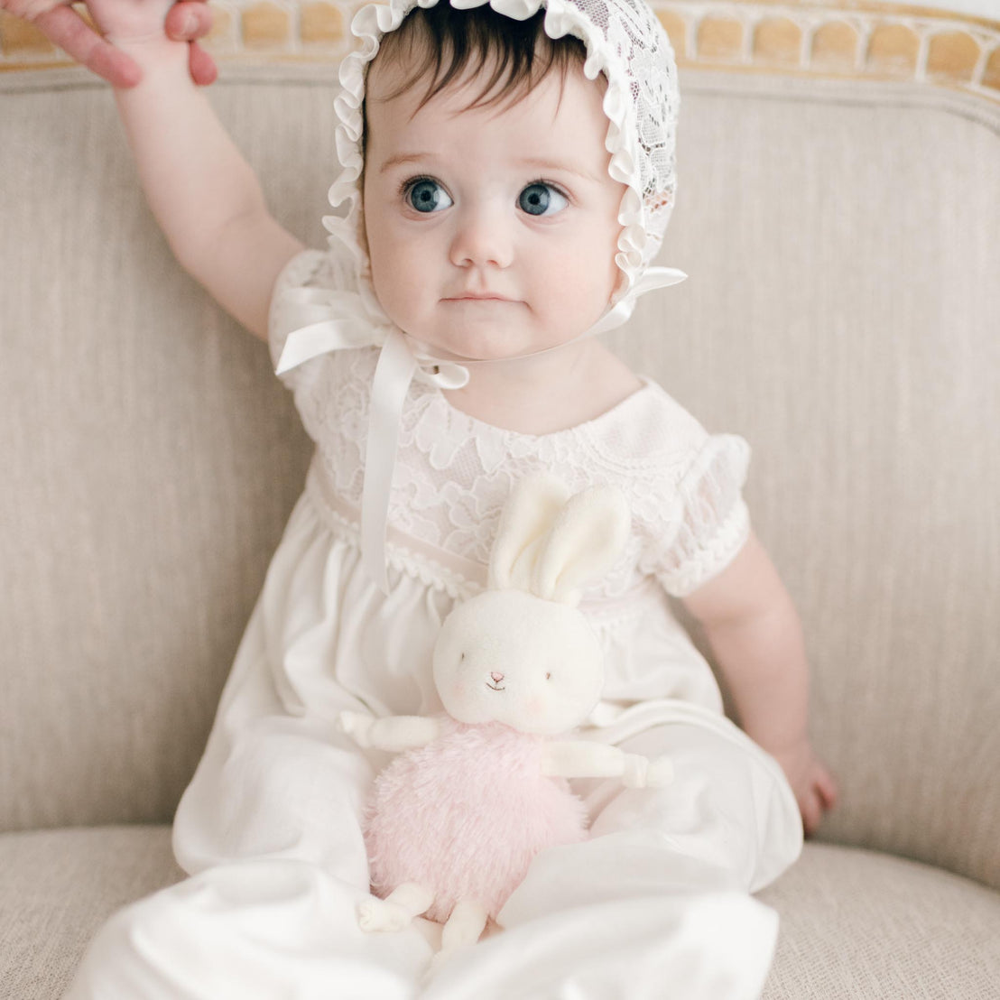 Baby girl in her baptism outfit with a plush bunny doll. Girl is sitting on couch holding her mother's hand.