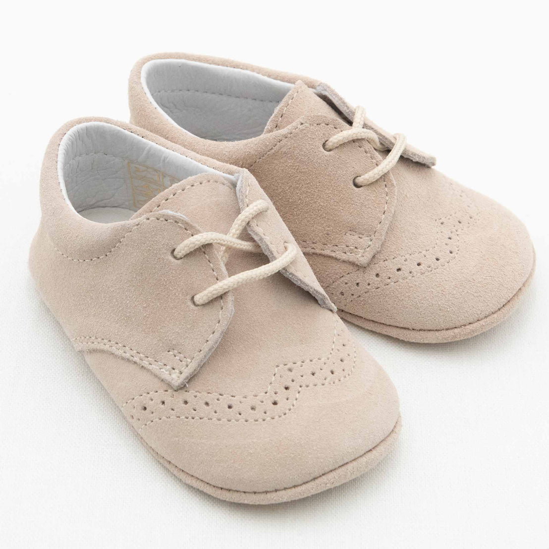 A pair of Ian Suede Shoes in light tan, crafted from soft suede material with white interiors. These baby boys' shoes feature decorative broguing on the toes and vamps, secured with white laces. The handmade suede shoes are placed on a white background.