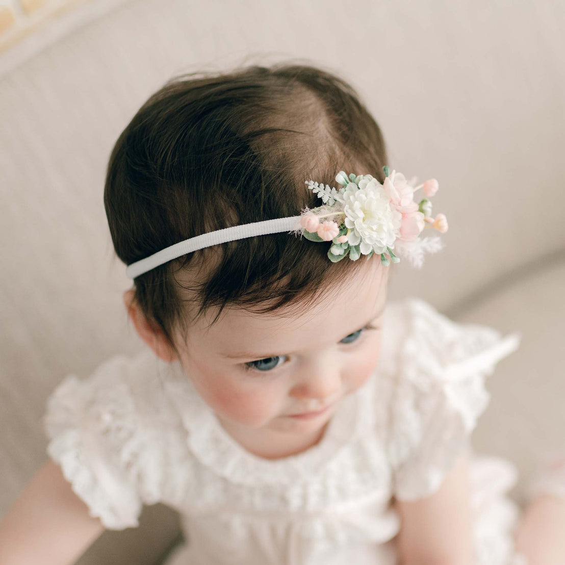 Top view of a baby girl wearing the Charlotte Flower Headband, featuring soft pink and white flowers. She stands near a linen chair, her head slightly turned and her eyes looking away.