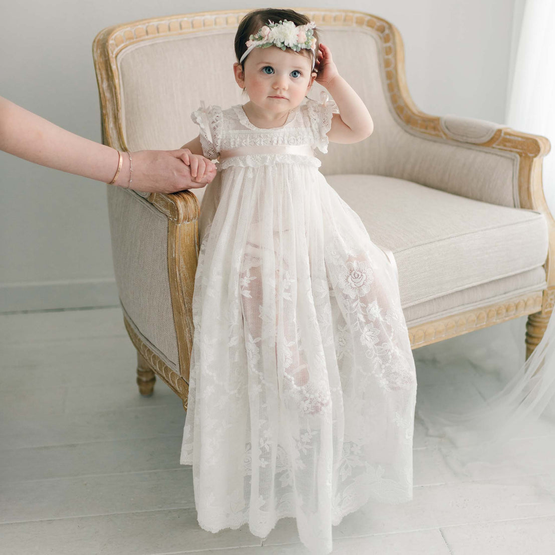 A toddler adorned in a handmade white dress paired with the Charlotte Convertible Skirt and a floral headband sits gracefully on a beige, upholstered chair. The child's hand is tenderly held by an adult's hand on the left side of the image. The softly lit room features neutral, minimalist décor, ideal for capturing a christening scene.