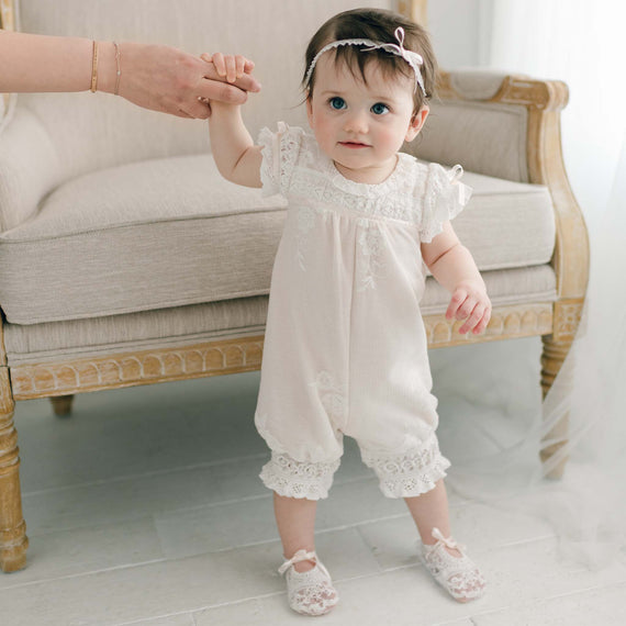 A baby girl with short dark hair, dressed in a light-colored, lace-trimmed Charlotte Romper and matching Charlotte Booties, stands while holding an adult's hand. The baby is also wearing a lace headband and is standing in front of a beige upholstered chair.