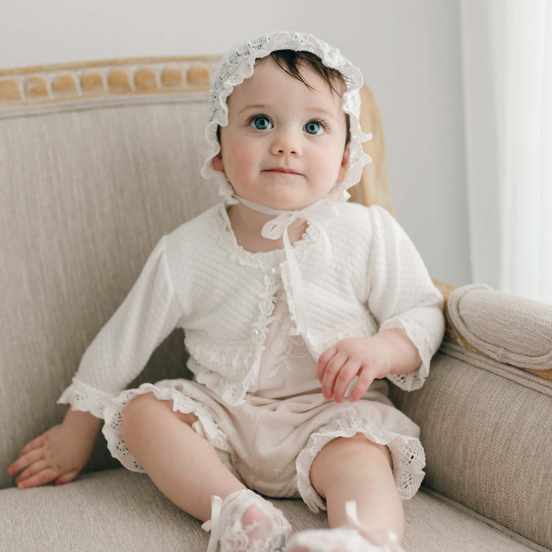 A baby with blue eyes is sitting on a beige upholstered chair. The baby is wearing the Charlotte Quilted Cotton Bonnet and is dressed in the Charlotte Romper and Quilted Cotton Sweater. The baby is looking slightly upward with a curious expression as natural light filters through the background.