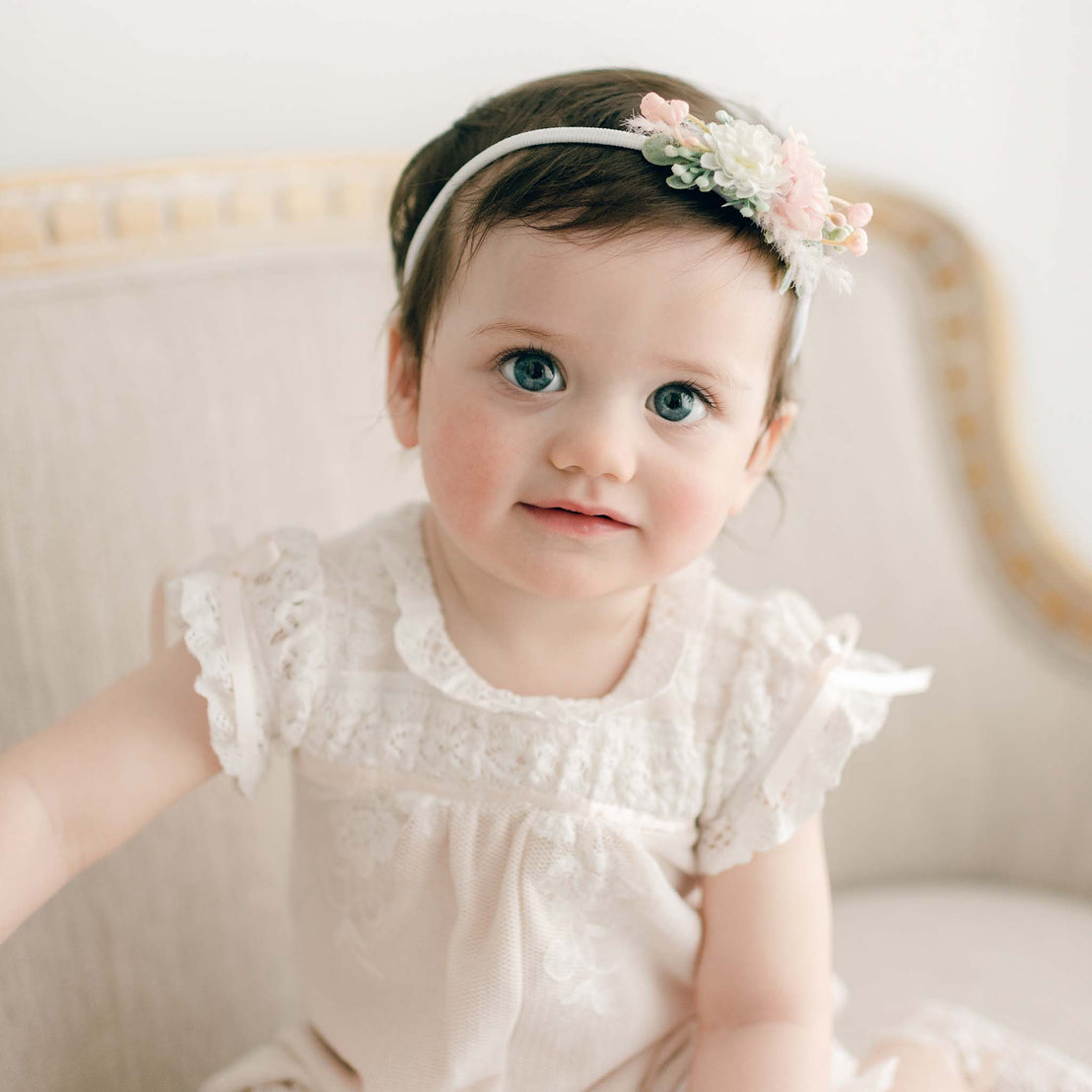 A baby girl wearing the Charlotte Romper and a floral headband. She is sitting in a beige upholstered chair.