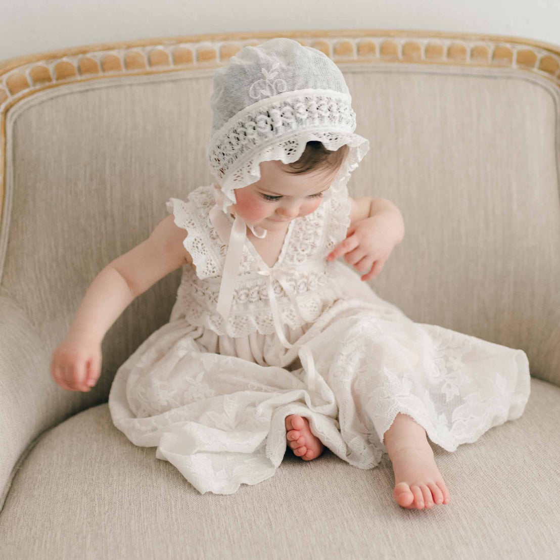 A toddler in a Charlotte Christening Gown & Bonnet sits on an elegant cream-colored chair, looking down thoughtfully. The background is a soft, beige and wood chair