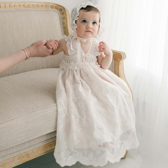 A toddler girl in a Charlotte Christening Gown & Bonnet sits on an elegant chair, looking curiously towards the camera with a slight frown. Her dark head is adorned with a matching lace bonnet.