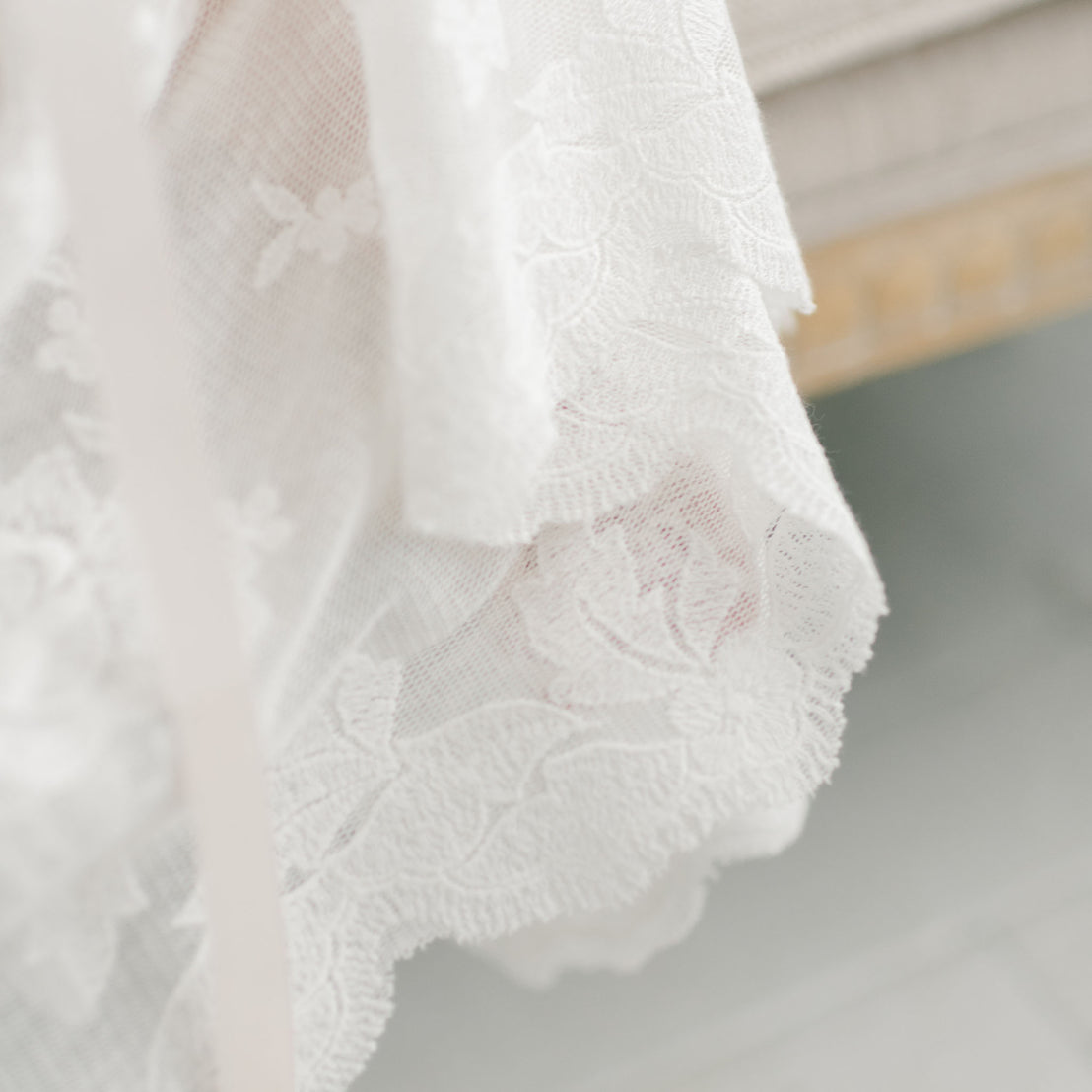 Close-up of the delicate white lace fabric with floral patterns from the Charlotte Convertible Skirt. The fabric, a key component of this versatile piece, features soft edges and a detailed texture. Handmade with care, it rests against an out-of-focus hint of beige upholstery in the background.