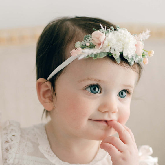 Close-up of a baby girl with blue eyes wearing the Charlotte Flower Headband with soft pink and white flowers. She is looking off to the side with her fingers gently touching her lips.