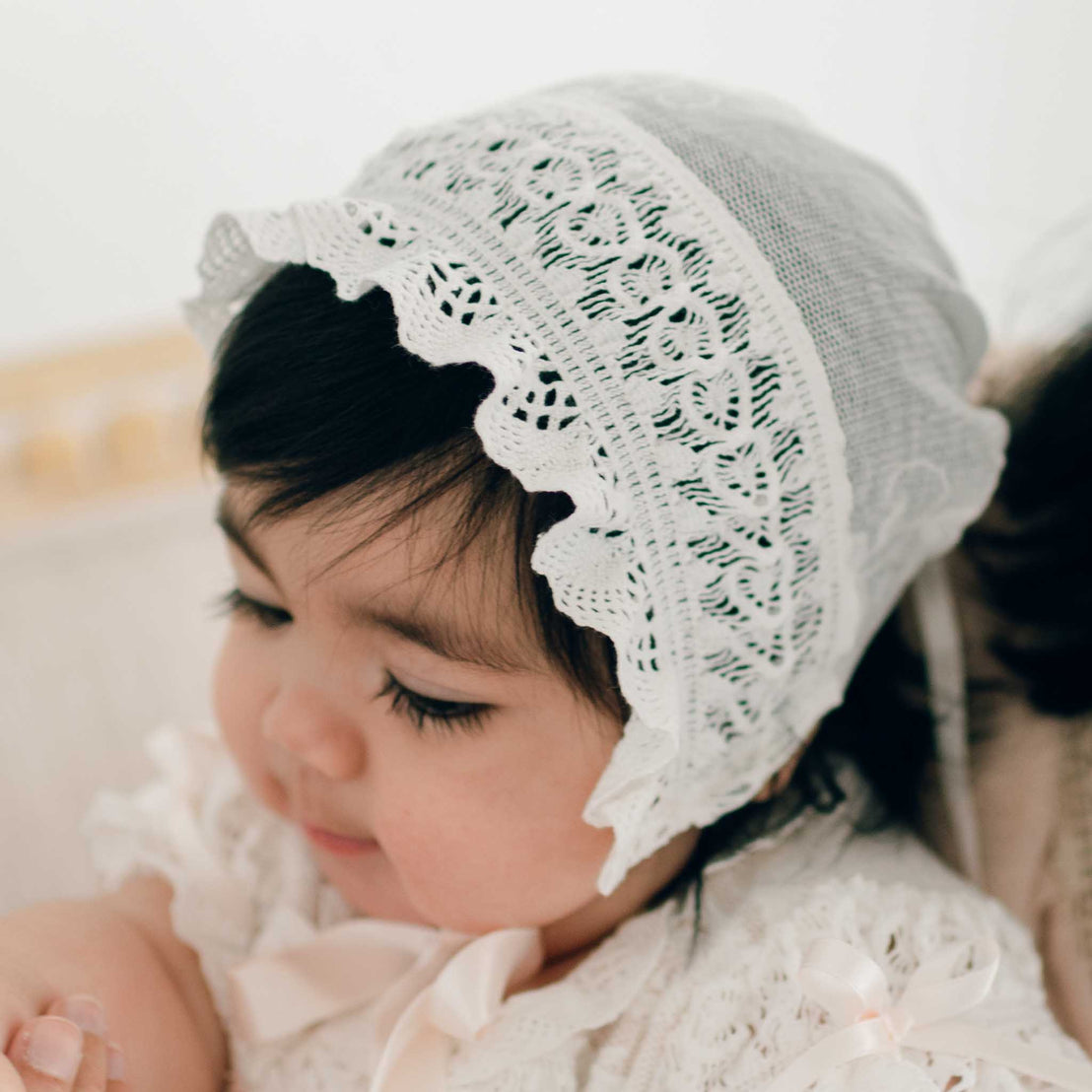 A baby wearing the Charlotte Christening Gown & Bonnet gazes downward, highlighting their long eyelashes and chubby cheeks. The soft lighting and neutral background accentuate the baby's peaceful expression, creating an ideal moment for a baptism gown photo or baby shower gift.