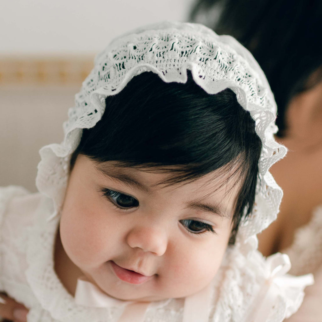 Dressed in the Charlotte Christening Gown & Bonnet, a baby gazes downward with a gentle expression. The softly blurred background highlights the baby's serene face, capturing a moment that becomes a cherished memory or the perfect baby shower gift.