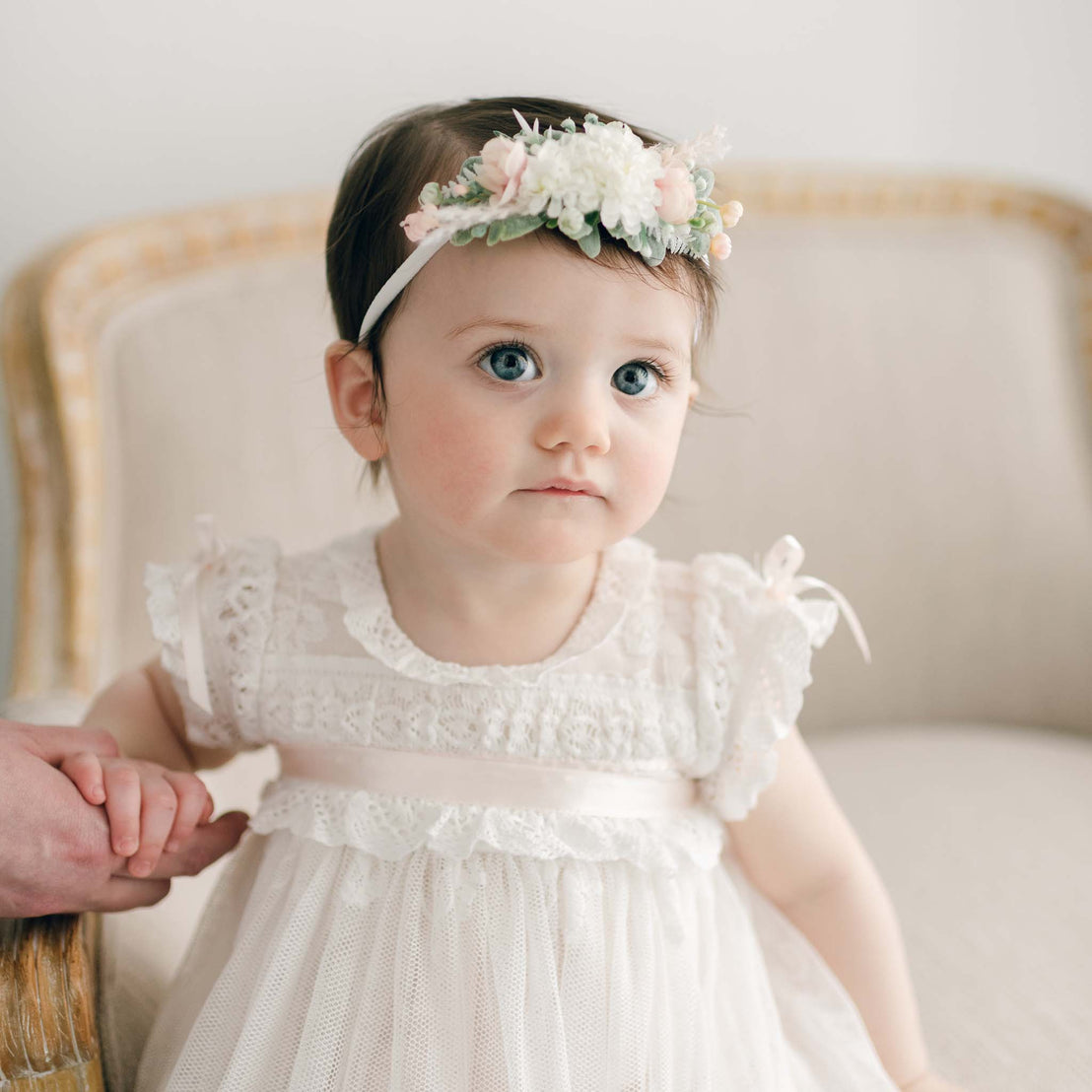 A baby girl wearing the Charlotte Convertible Skirt & Romper Set and a floral headband. Someone is holding her right hand. In the background is a beige upholstered chair.