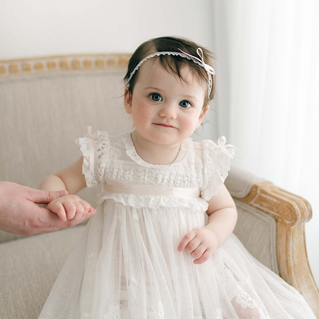 A toddler with short brown hair, dressed in the elegant Charlotte Convertible Skirt paired with a handmade white lace dress and a matching headband adorned with a small bow, sits on a cream-colored armchair. An adult hand supports the child's left hand. The softly lit background, featuring white curtains, evokes an air of christening grace.
