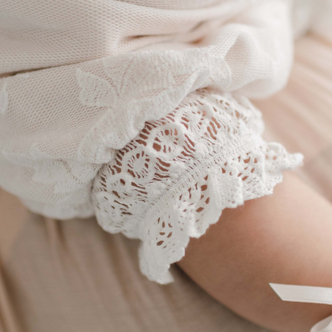 Close-up of a baby's leg dressed in the Charlotte Romper. It is a part of the Charlotte Convertible Skirt & Romper Set. The baby is resting on light beige fabric, enhancing the image's softness with its delicate embroidered cotton lace details.