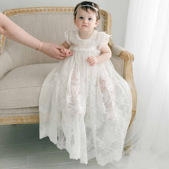 A toddler in a Charlotte Convertible Skirt sits on a cream-colored armchair, adorned with a matching headband and gazing slightly to the side while holding an adult's hand. The background features sheer white curtains, enhancing the soft ambiance of the scene.