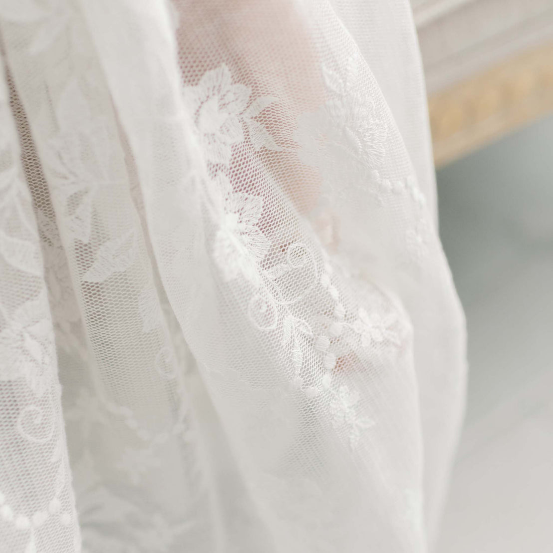 Close-up of the Charlotte Convertible Skirt's delicate white lace fabric featuring an intricate floral pattern, making it ideal for a christening skirt. The lace is softly draped, showcasing fine details of flowers and leaves within its handmade textile design. The background is slightly blurred, emphasizing the fabric's texture and craftsmanship.