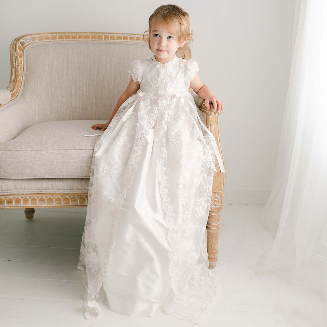Baby girl making funny face wearing a full-length Penelope christening gown, made of super-soft silk and luxurious floral netting.