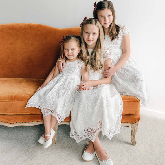 Three young girls in Grace Girls Lace Holiday Dresses sit on an orange velvet couch. Their outfits, made from a delicate blend of silk and cotton, bring a touch of elegance to the scene. They smile warmly, with the older two hugging the youngest, all surrounded by a simple, bright interior.