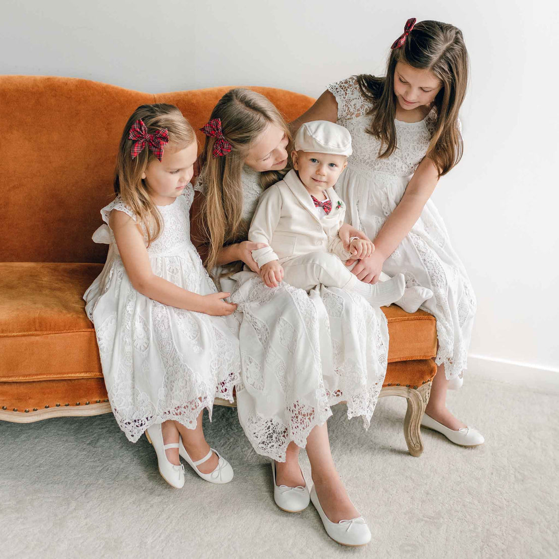 A woman and three children in white lace outfits sit on an orange sofa. The children are wearing matching Plaid Bow Hair Clips in their hair, adding a charming touch, while the baby boy wears a white hat and suit. They're smiling and looking content.