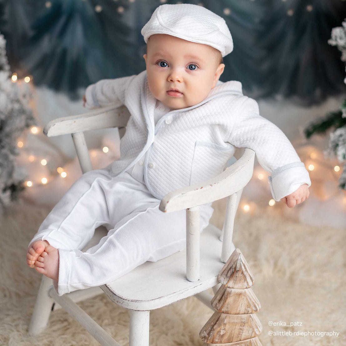 A baby dressed in a Harrison 3-Piece Suit sitting on a small vintage wooden chair with a backdrop of twinkling lights, upscale baptism decorations.