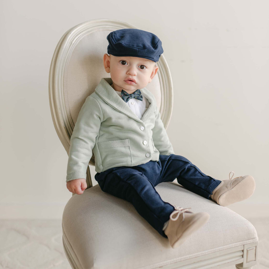 A baby dressed in the Cole 3-Piece Suit, which includes a light green cardigan, white shirt, navy bow tie, and matching navy pants, sits on a cream chair. Handmade in the USA from French Terry cotton, they accessorize with a navy flat cap and light tan shoes against a neutral backdrop, making it an ideal outfit for any holiday party.