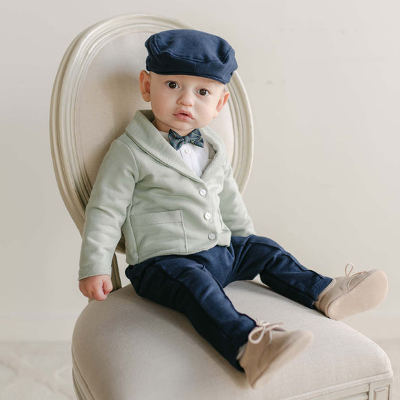 A baby dressed in the Cole 3-Piece Suit, which includes a light green cardigan, white shirt, navy bow tie, and matching navy pants, sits on a cream chair. Handmade in the USA from French Terry cotton, they accessorize with a navy flat cap and light tan shoes against a neutral backdrop, making it an ideal outfit for any holiday party.