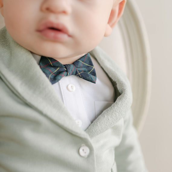 A baby dressed in a mint green cardigan and white shirt sports the Cole Bow Tie & Boutonniere Set, featuring a handmade dark bow tie with a subtle pattern. The focus is on the clothing and accessories, with the baby's face partially visible against a softly blurred background.