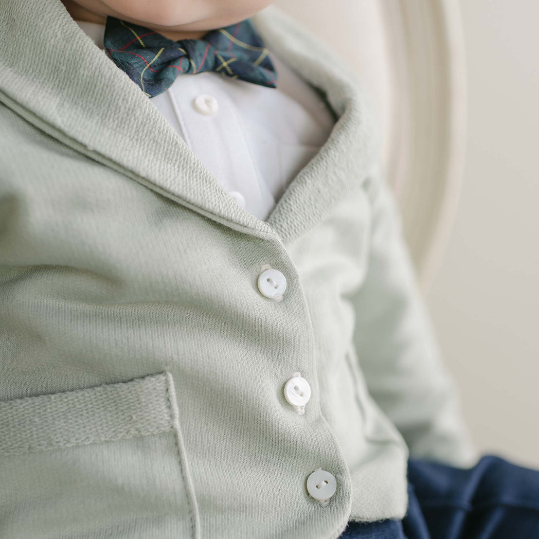 Close-up of a child wearing the Cole 3-Piece Suit, featuring a light green cardigan crafted from French Terry cotton with white buttons over a white shirt. The ensemble is completed with a dark, colorful patterned bow tie, making it ideal for a holiday party. Handmade in the USA, this outfit stands out against the neutral background.