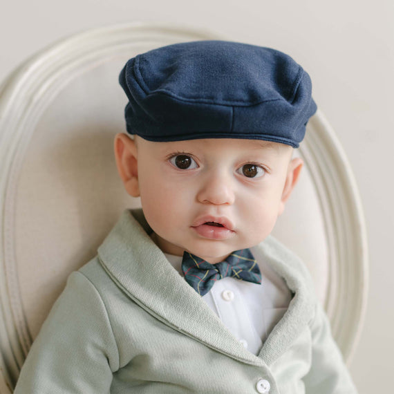 A baby wearing a Cole Newsboy Cap, along with a light green french terry cotton sweater and a plaid bow tie, sits on a cream-colored chair, looking curiously at the camera.