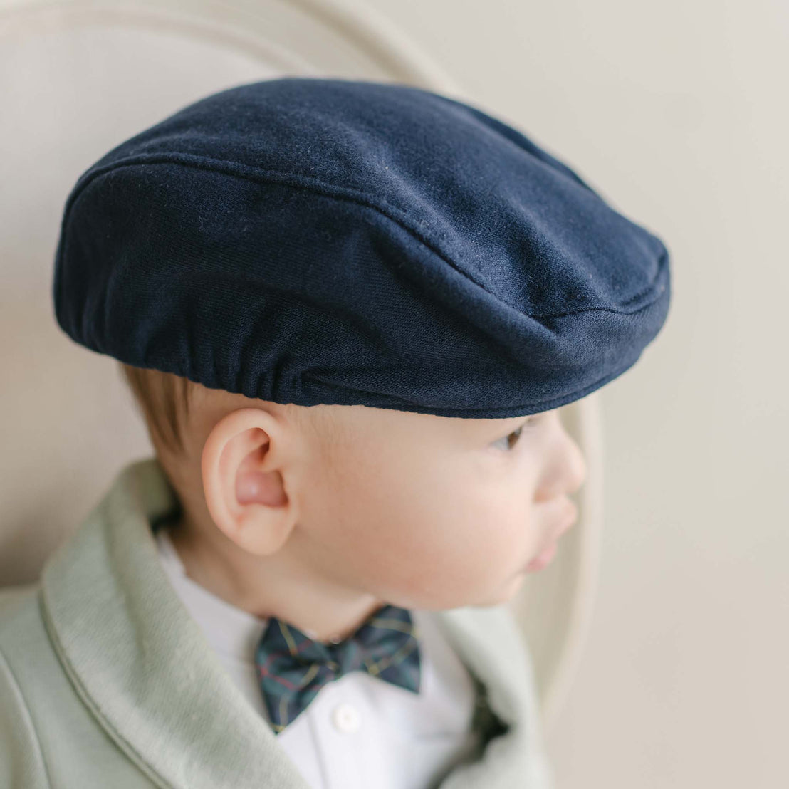 A young child wearing a Cole Newsboy Cap in navy blue and a plaid bow tie is seated in a soft chair. Dressed in an outfit made of light-colored French terry cotton, the child gazes to the side. The neutral background contributes to the image's soft, classic feel.