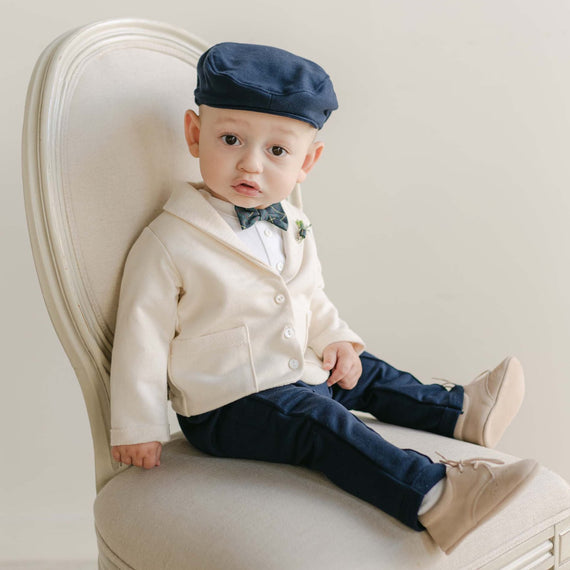 A baby, dressed in the Cole 3-Piece Suit which includes a cream-colored French Terry cotton jacket, navy pants, and complemented by a blue bow tie and beige shoes, sits on a white chair. The ensemble is completed with a handmade flat cap from the USA. The baby gazes directly at the camera against a neutral backdrop, creating an ideal scene for a holiday party photo.