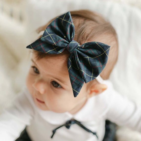 A baby wearing a white shirt and the Colette Bow Headband sits on a light-colored blanket, looking curiously to the side. The stylish, blue plaid cotton headband adds an adorable touch.