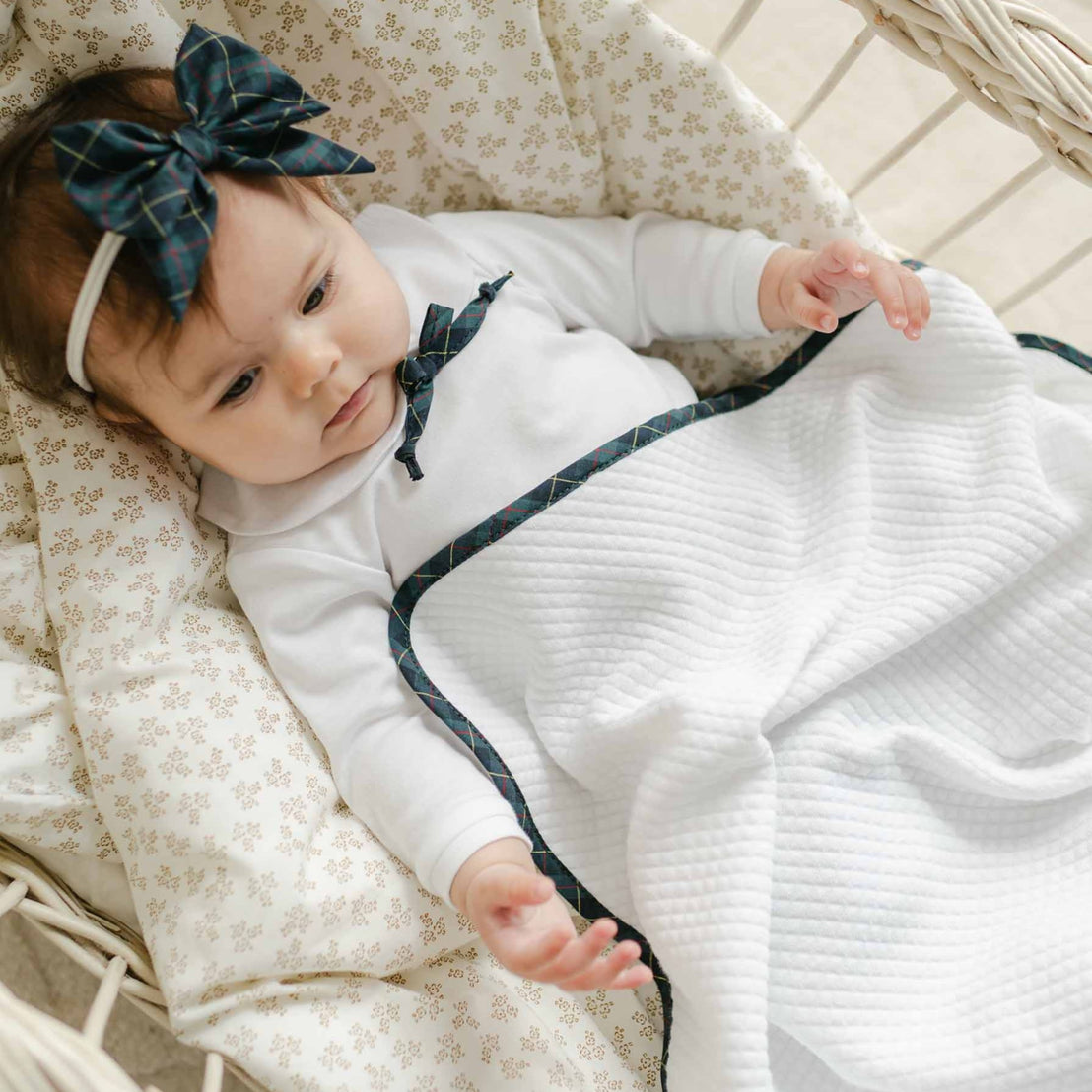 A baby lies in a wicker bassinet, dressed in a white outfit with blue trim and a matching blue headband with a bow. The little one is snuggled under the Cole Personalized Blanket, resting on a patterned cushion.