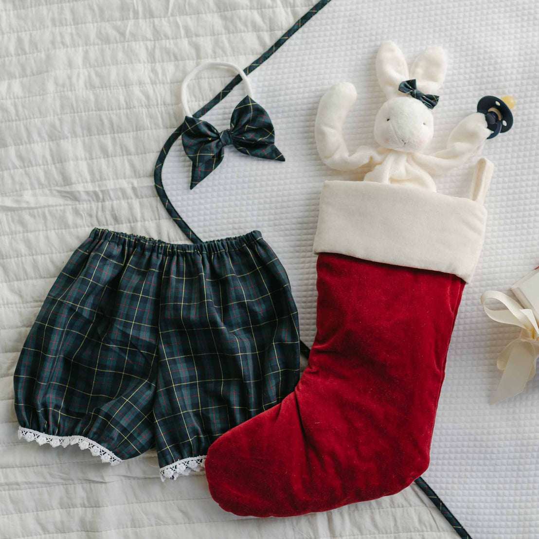 A red Christmas stocking with a plush rabbit inside rests on a quilted surface. Nearby, the Colette Bloomer Set featuring a blue plaid onesie and lace-trimmed shorts is accompanied by a headband with a bow. A baby pacifier is also visible next to the rabbit, ready for festive holiday events.