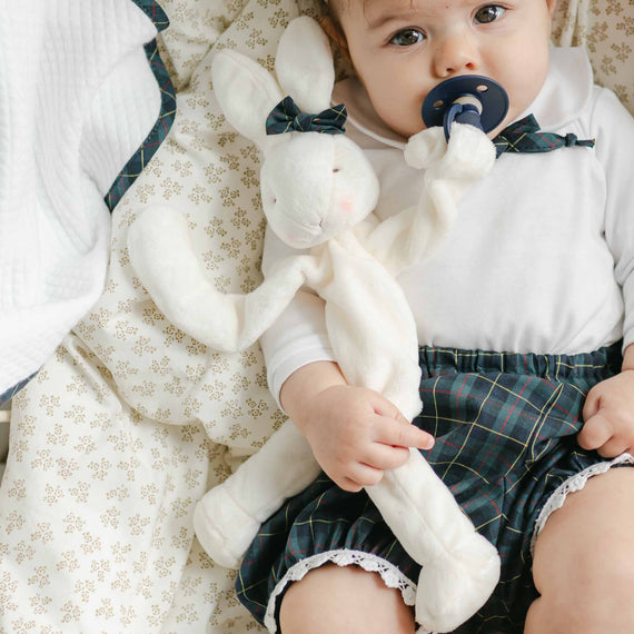 A baby snuggles with a Cole Silly Bunny Buddy, securely attaching their pacifier to their soft velour shirt, and looks adorable in a safe-to-wear ensemble of a white shirt and plaid shorts with lace trim.