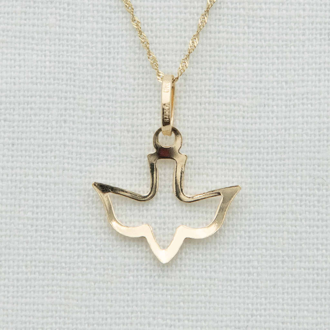 A 14k Gold Dove Charm with Chain featuring a pendant in the shape of an abstract dove. The solid gold pendant hangs from a twisted gold chain, making it the perfect accessory for a christening outfit. The background is a light, textured fabric.