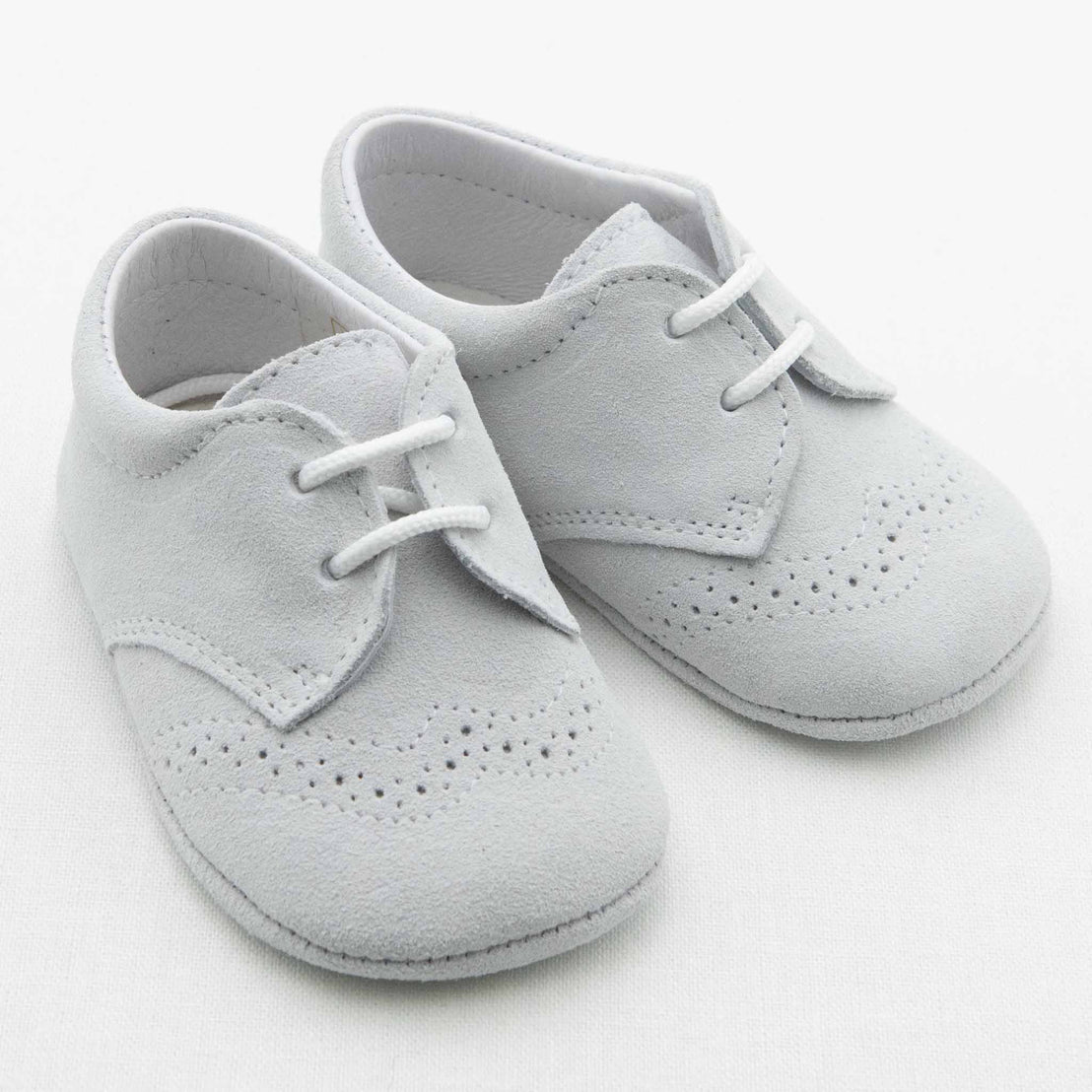 A pair of Dove Grey Suede Shoes for baby boys crafted from soft suede material. Handmade in Spain, these shoes boast a classic design with decorative brogue perforations adorning the front and edges. They feature white laces and a low heel, showcased on a plain white background.