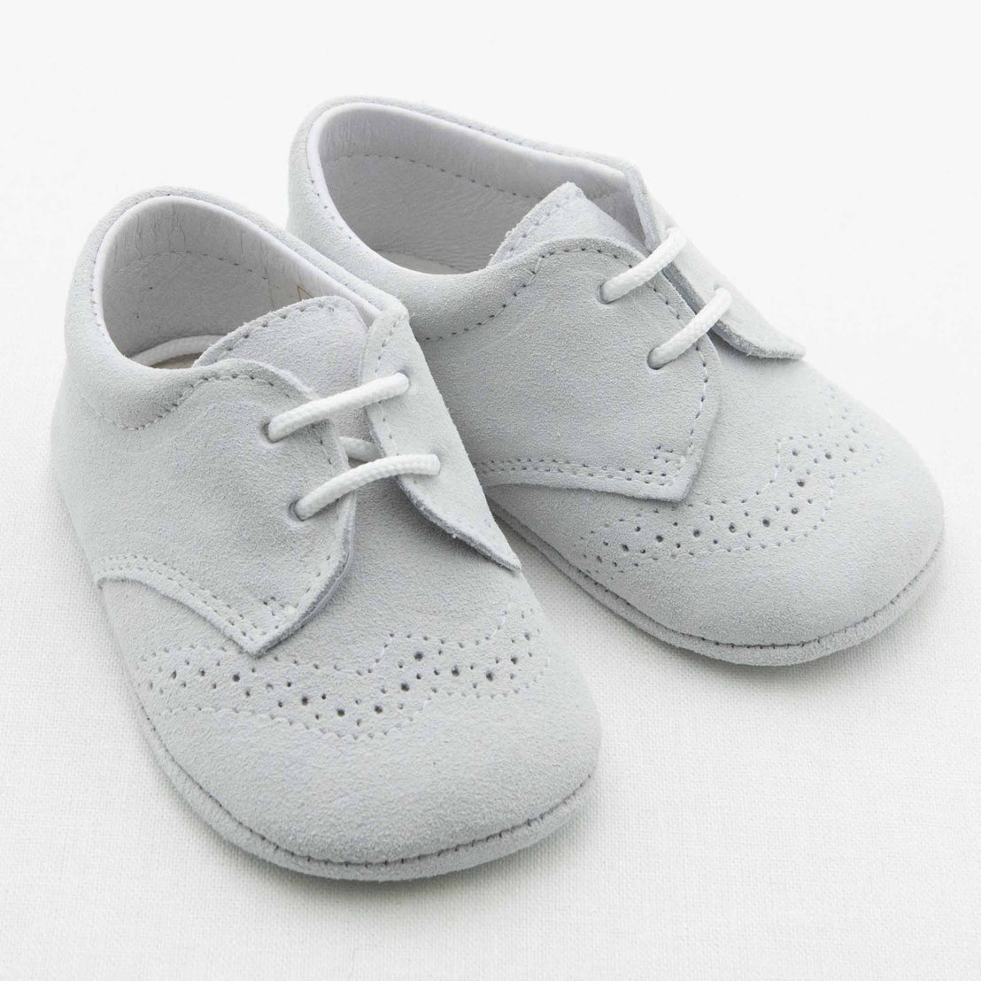 A pair of Boys Suede Shoes made of soft, light grey fabric with laces. Part of the Baby Beau & Belle collection, these charming handmade baby shoes feature decorative perforations along the edges and a slightly pointed toe. The beautifully crafted shoes are showcased on a white background.