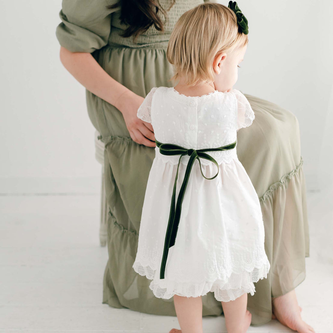 A toddler in the Emily Dress & Bloomers with a Green Sash stands in front of a seated woman, who adjusts the child's dress for a christening. The setting is minimalist with.