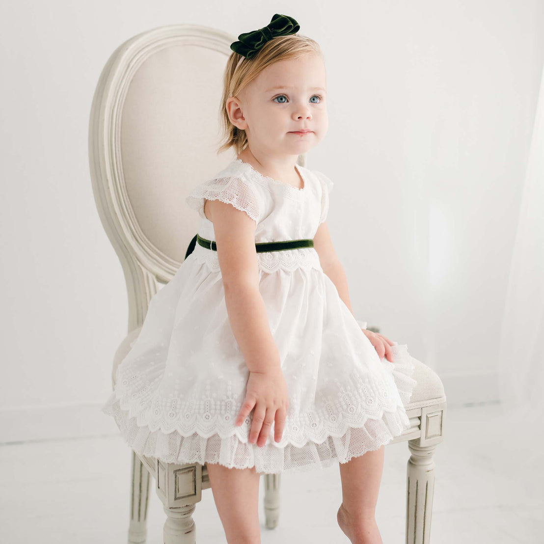 A young girl in an Emily Dress & Bloomers with a Green Sash sits on an upscale chair, looking off to the side, in a bright, minimalist room.