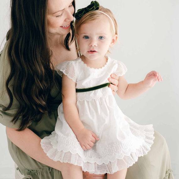 A mother, smiling gently, holds her young daughter, dressed in the Emily Dress & Bloomers with a green sash and headband during her baptism. They are both seated in a softly lit.