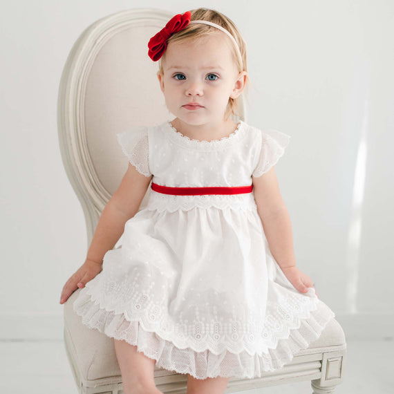 A toddler with fair skin and light hair sits on an upscale white chair, wearing the Emily Dress & Bloomers with lace detailing and a red belt for her baptism.